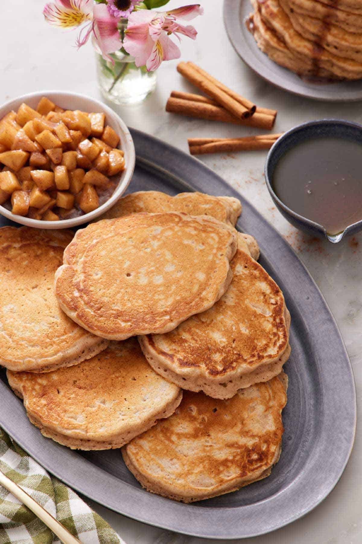A platter of apple pancakes with a bowl of cooked apples. A bowl of syrup and cinnamon sticks beside the platter.