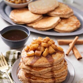 Pinterest graphic of a stack of apple pancakes topped with apples and syrup. More pancakes and cooked apples in the background with cinnamon sticks and syrup in between.