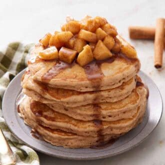 A plate with a stack of apple pancakes topped with cooked apples and syrup. Cinnamon sticks in the background and a fork beside the plate.