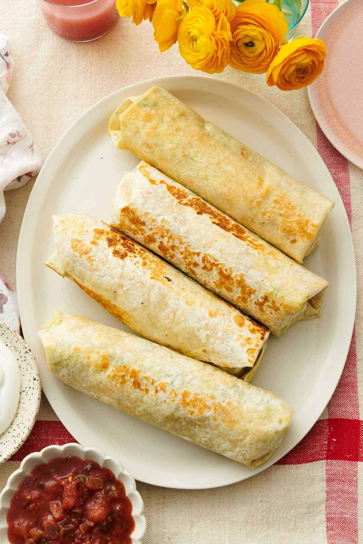 Overhead view of breakfast burritos on a white plater. A bowl of salsa, sour cream, and flowers on the side.