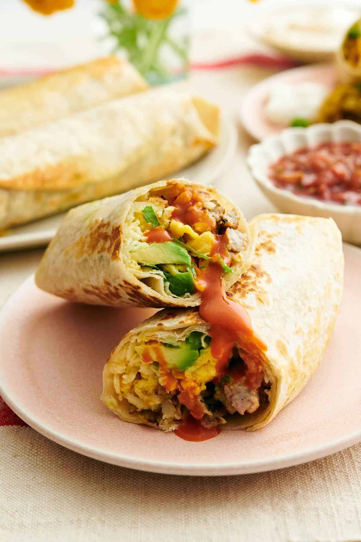 A plate with a breakfast burrito cut in half, one half stacked on top of the other, showing the interior. More burritos in the background and a bowl of salsa.