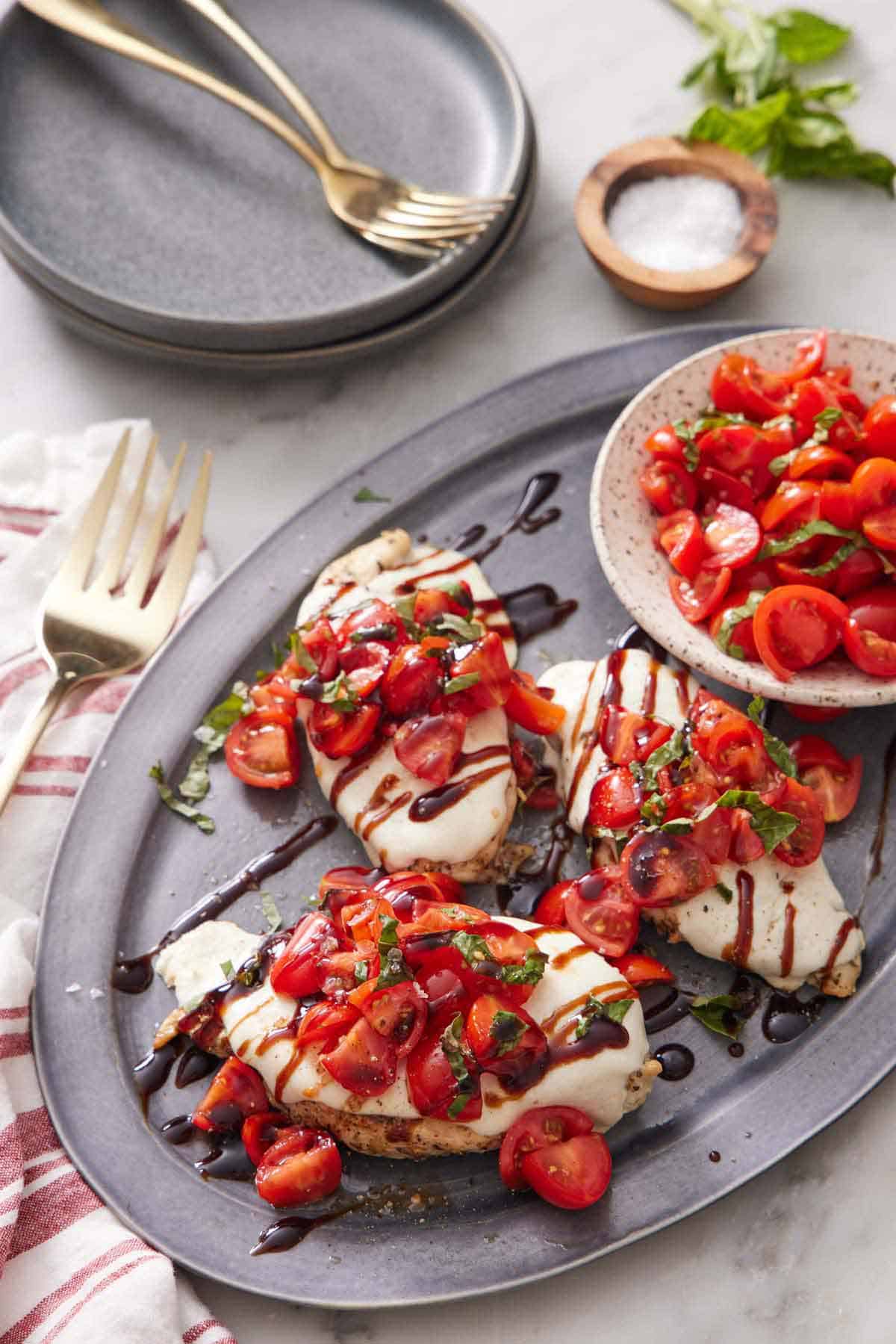 A platter with three bruschetta chicken with a bowl of tomatoes. Each chicken topped with balsamic glaze and tomatoes on top.
