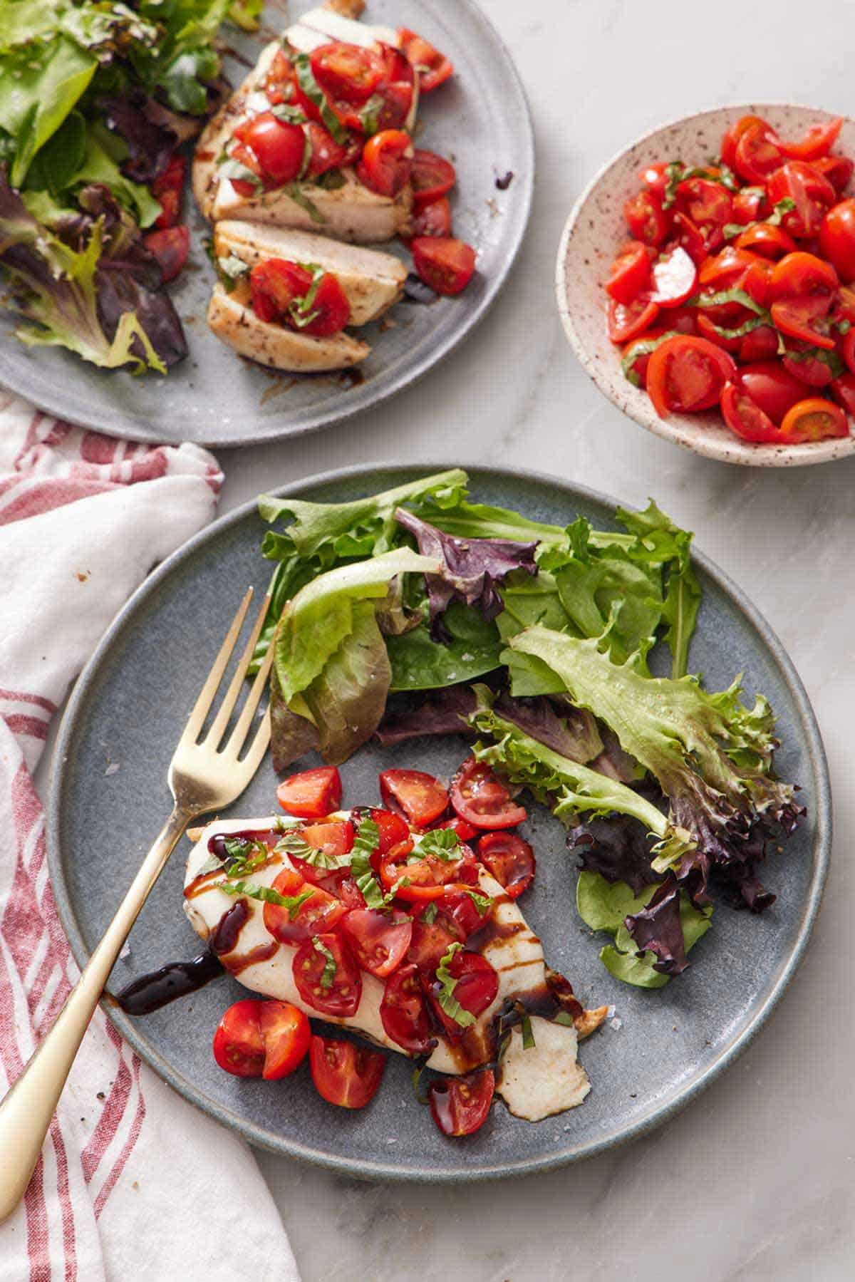An overhead view of a plate of bruschetta chicken and mixed greens with a fork. Another plate in the background along with a bowl of seasoned tomatoes.