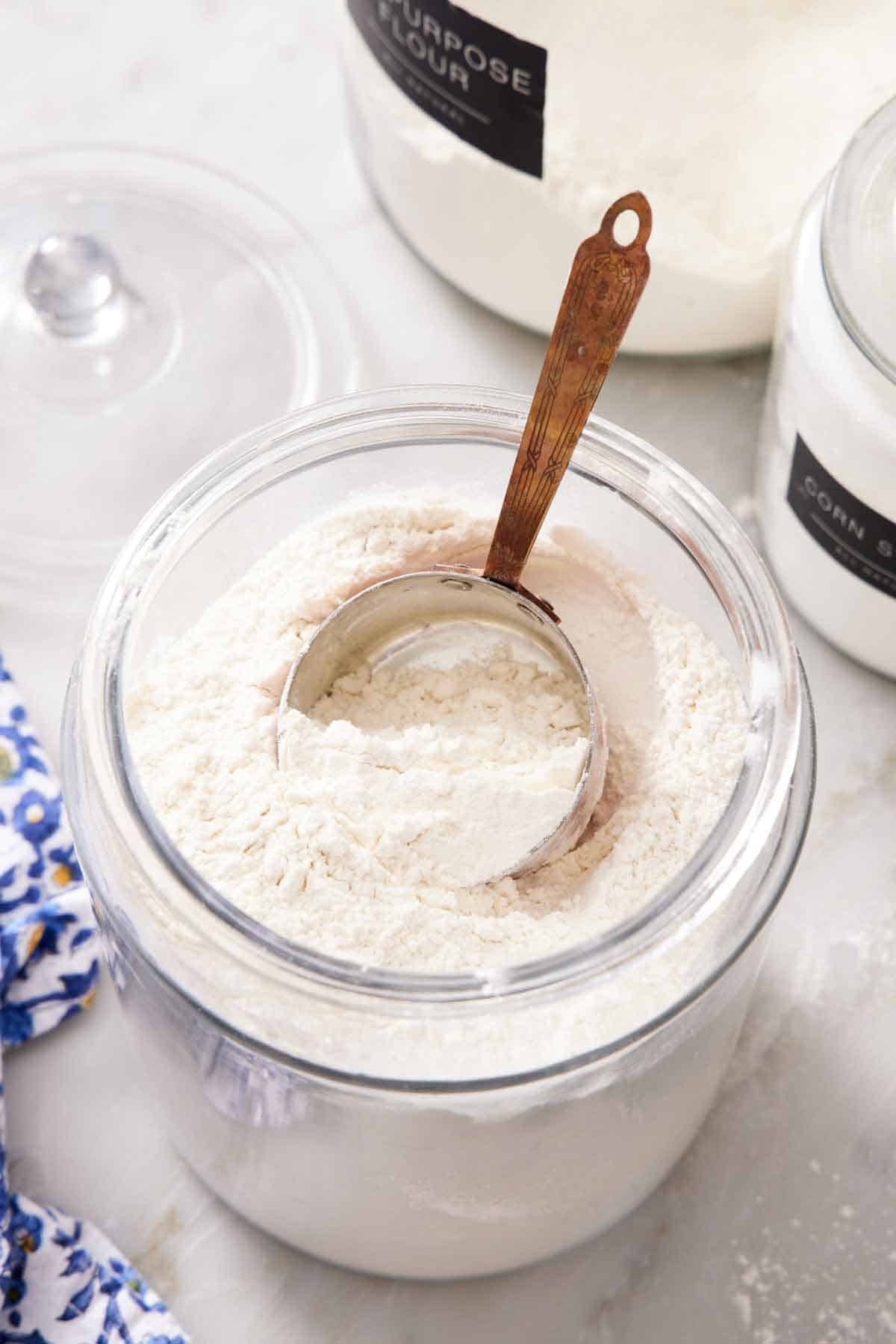 A large jar of cake flour substitute with a measuring cup inside. Two more jars of flours in the background.