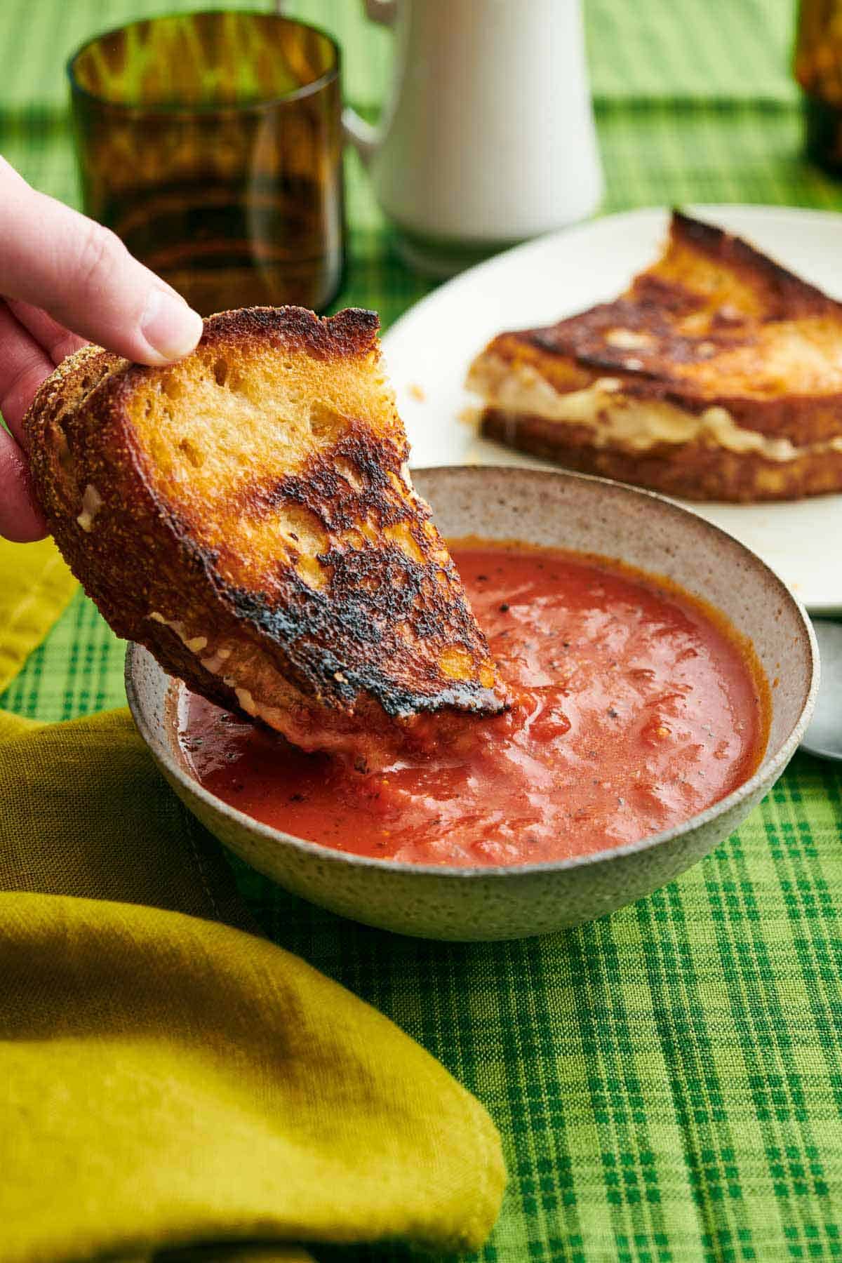 Half of a grilled cheese dipped into a bowl of tomato soup. A plate with the other half of the grilled cheese in the background.