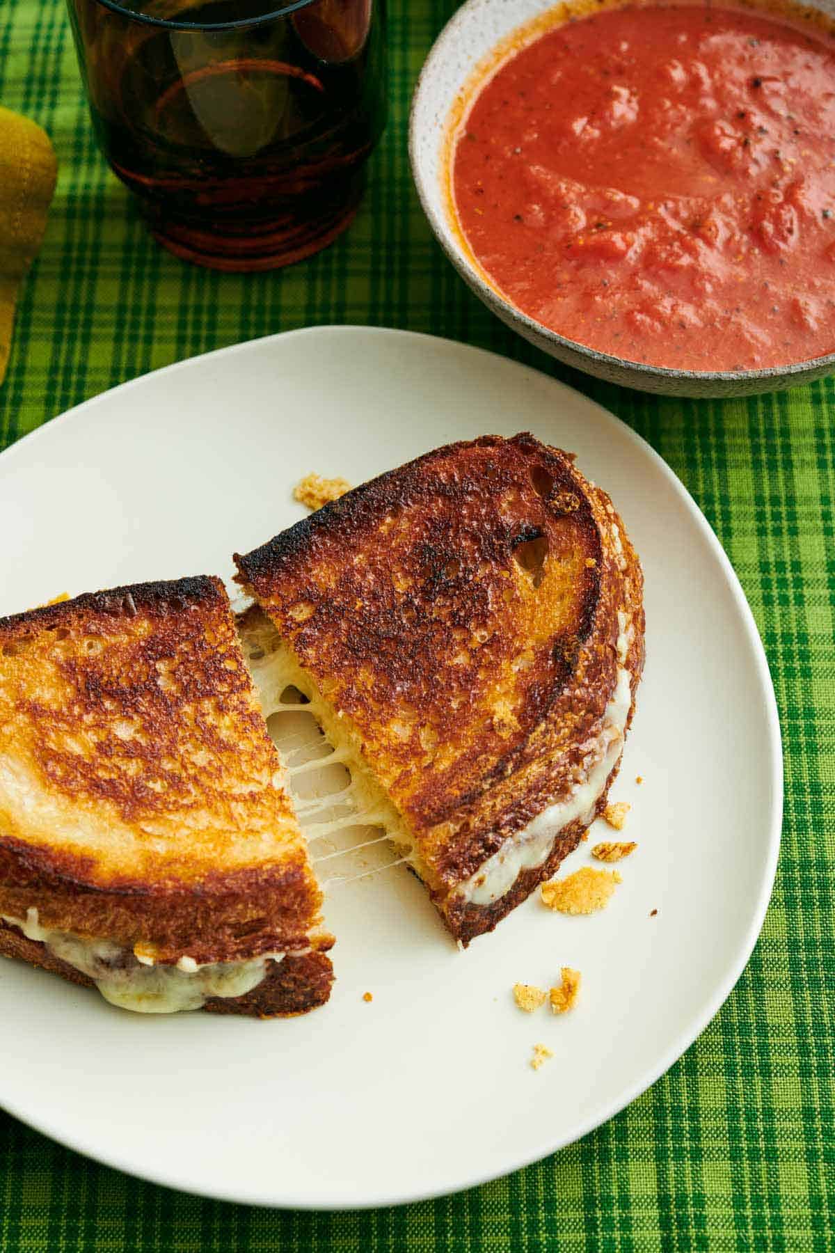 A plate with a grilled cheese cut in half and slightly pulled apart. A bowl of tomato soup beside it.