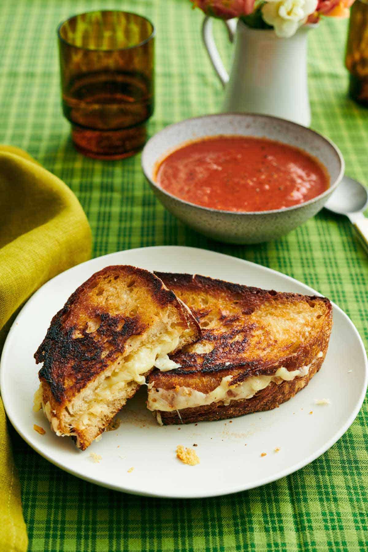 A plate with a grilled cheese cut in half, one half leaning up against the other. Bowl of tomato soup in the back along with a drink.