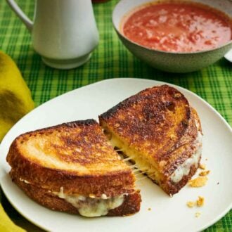 Pinterest graphic of a plate with a grilled cheese cut in half and slightly pulled apart. A bowl of tomato soup and vase of flowers in the back.