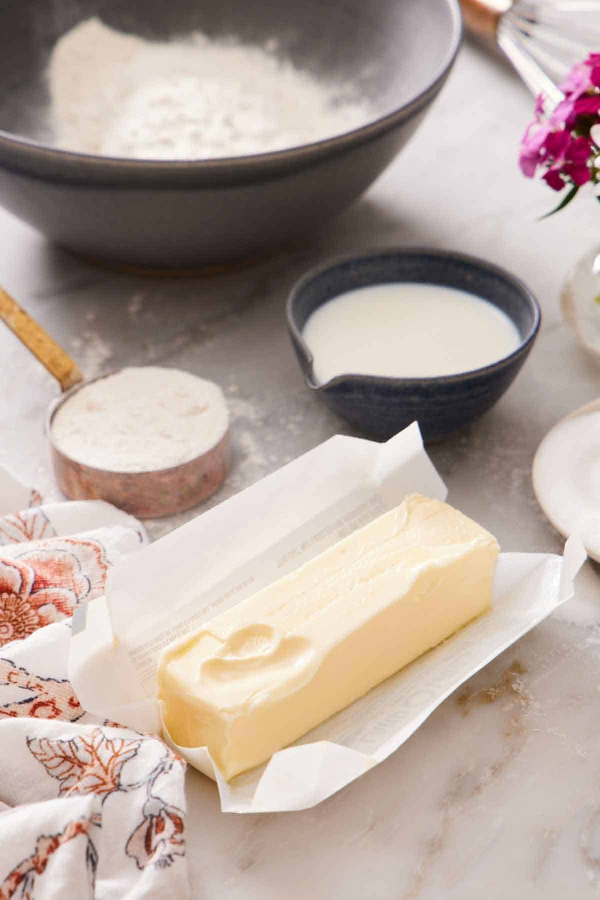 A stick of softened butter in an opened wrapper. A bowl and measuring cup of flour and dairy in the background.