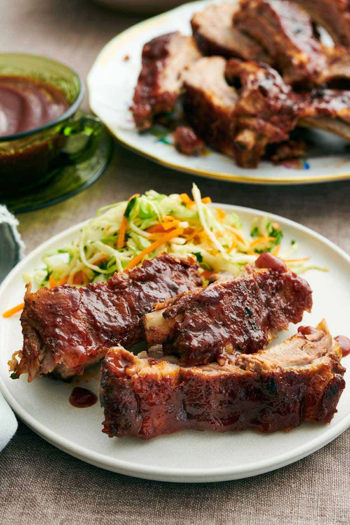 A plate with Instant Pot ribs with vegetable slaw. A platter with more ribs and a bowl of barbecue sauce in the back.