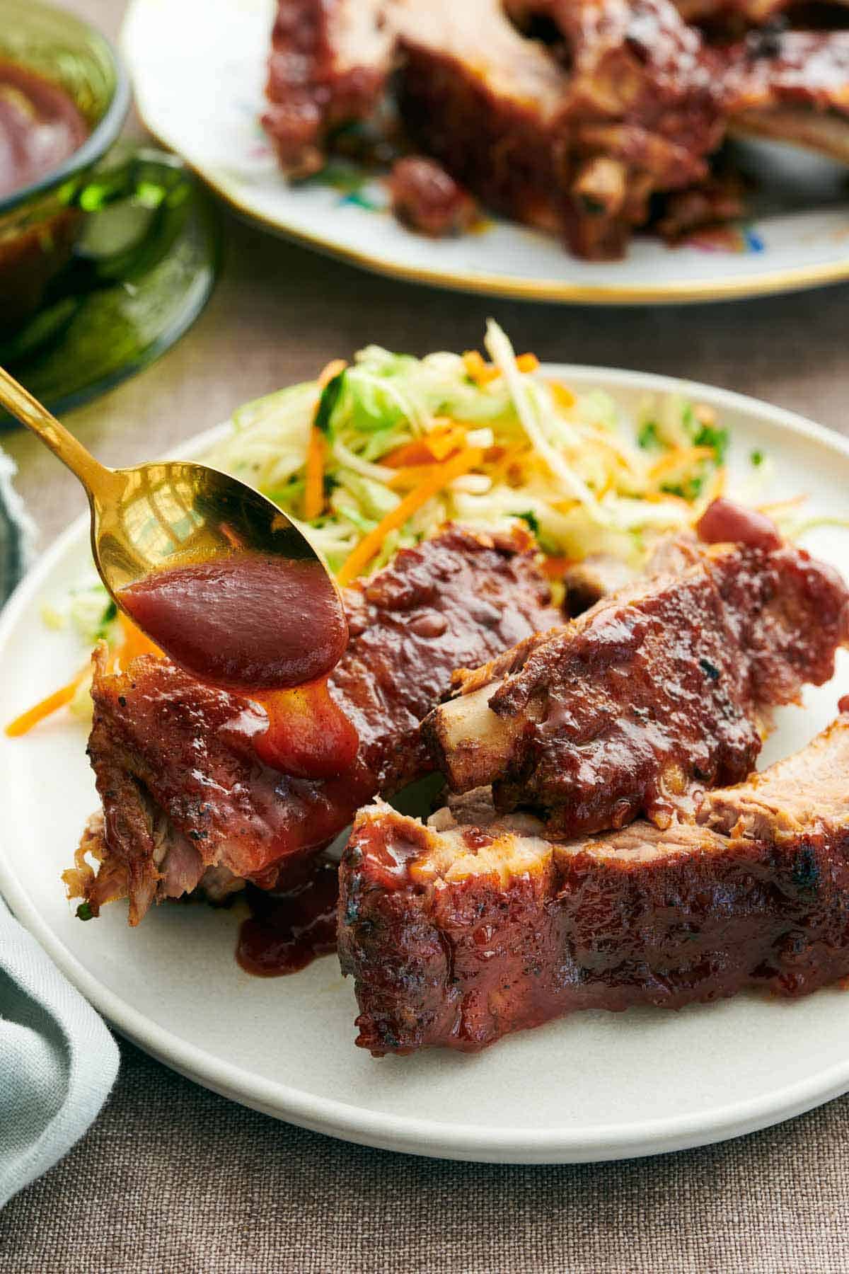 A spoonful of barbecue sauce drizzled over Instant Pot ribs on a plate. Vegetable slaw on the plate with the ribs and more ribs in the background.