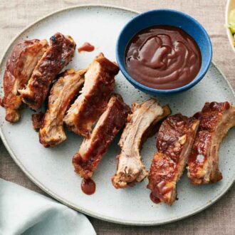 Overhead view of a platter of Instant Pot ribs with a bowl of barbecue sauce. A bowl of vegetable slaw on the side.