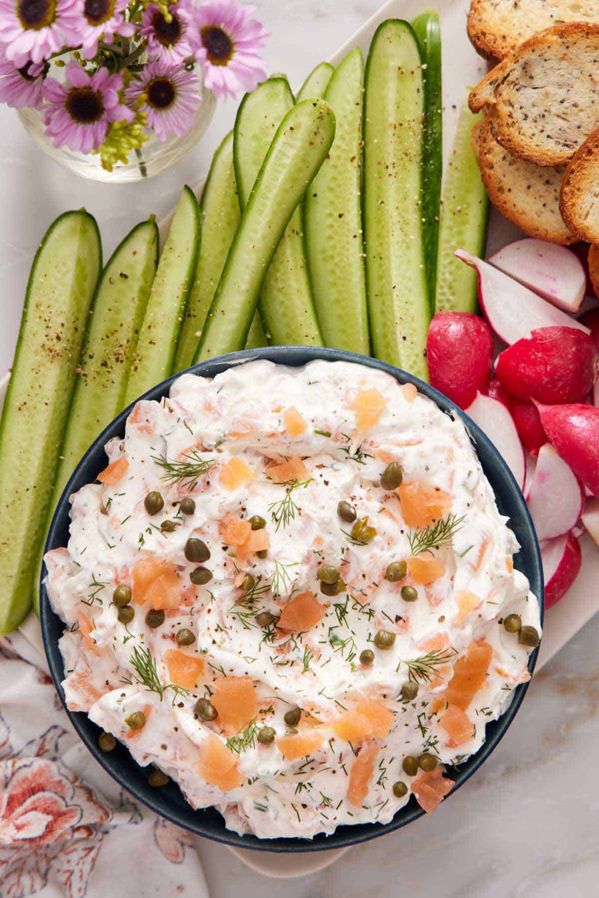 A bowl of smoked salmon dip topped with fresh dill and capers. Sliced cucumbers, radishes, and bread on the the side.