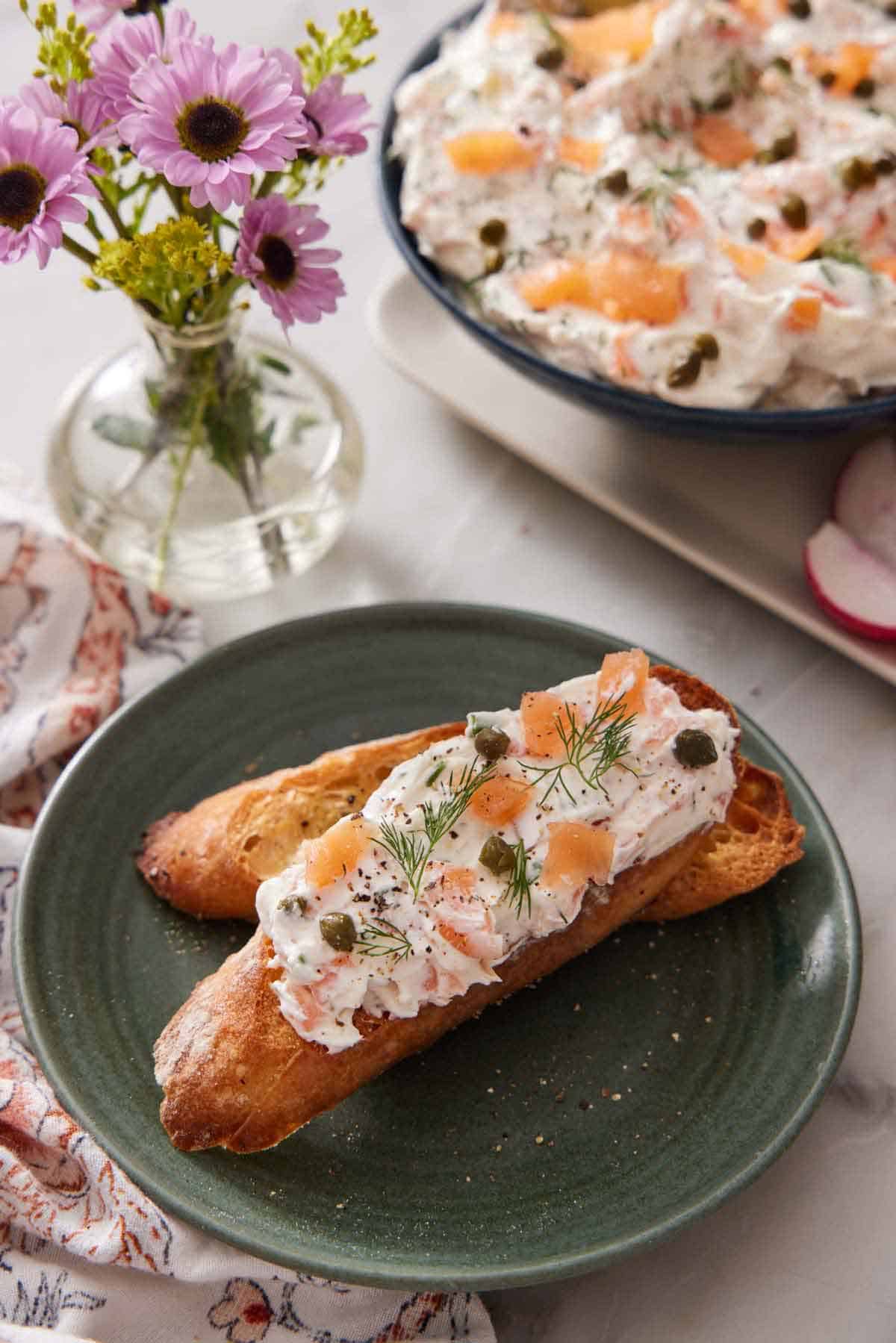 A toasted piece of bread with creamy smoked salmon dip, topped with capers and dill. A bowl of more dip in the background along with a vase of flowers.