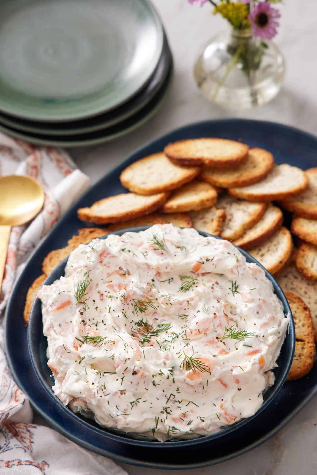 A bowl of smoked salmon dip topped with fresh dill on a platter with crackers. A vase in the background with a stack of plates.