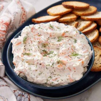 A bowl of smoked salmon dip topped with fresh dill on a platter with crackers. a linen napkin on the side.
