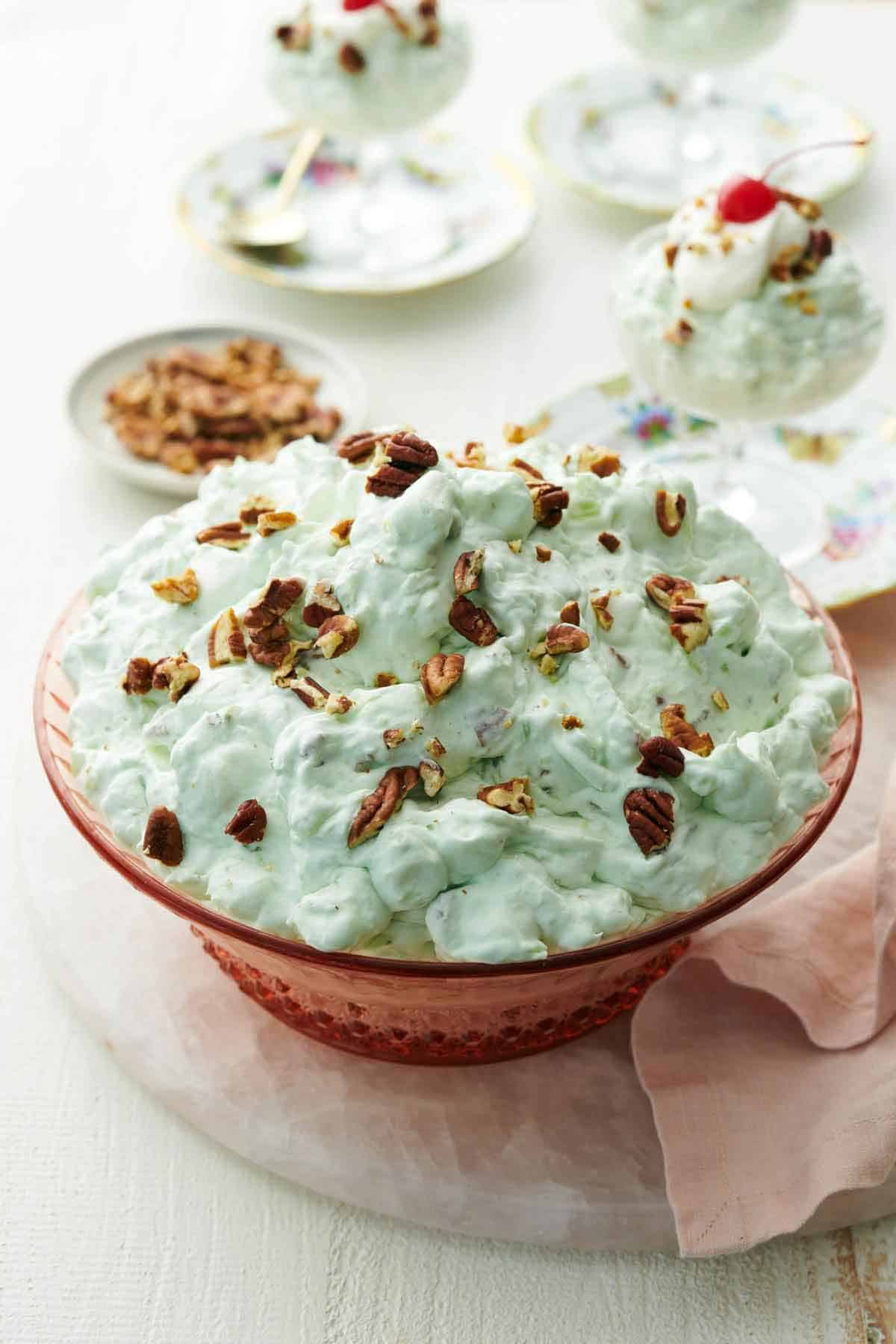 A serving bowl of Watergate salad topped with chopped pecans. Three servings in dessert bowls in the background.