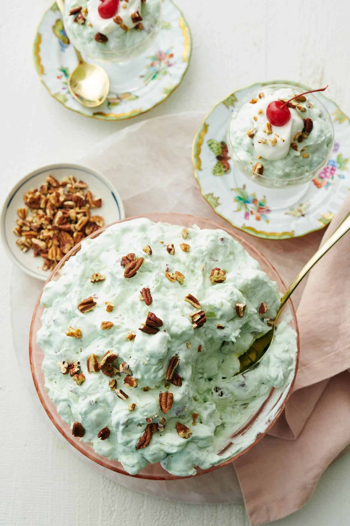 Overhead view of a bowl of Watergate salad with chopped pecans on top with a spoon tucked in. A bowl of pecans and two servings plated on the side.