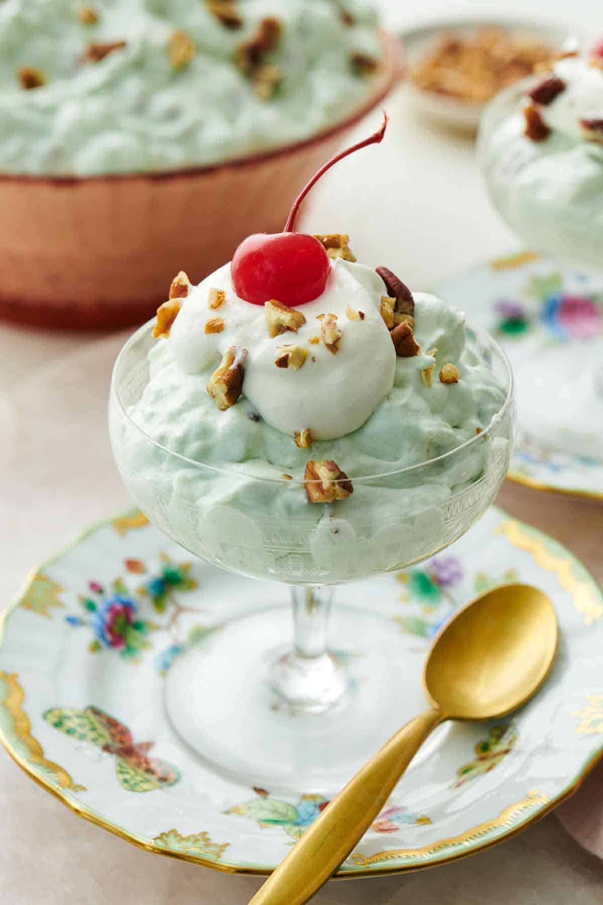 A dessert bowl with Watergate salad topped with whipped cream, pecans, and a maraschino cherry. A spoon beside it.