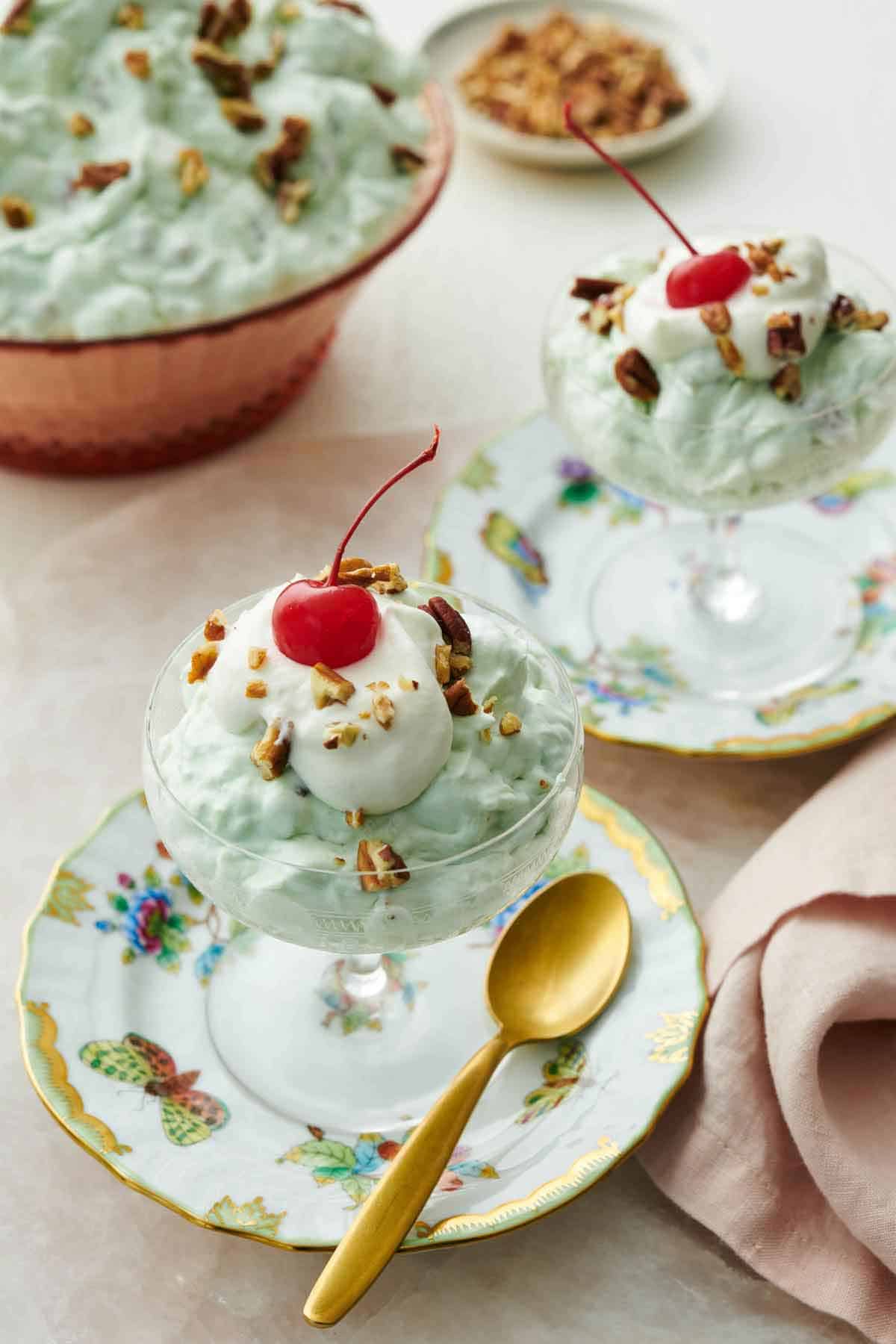 Two dessert bowls on plates with Watergate salad topped with whipped cream, pecans, and a maraschino cherry. A bowl of more Watergate salad in the background.