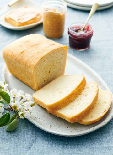 A platter with a loaf of white bread with three slices cut in front. Peanut butter and jam in the background along with a slice of bread with peanut butter.