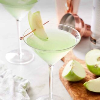 Pinterest graphic of glasses of apple martini with sliced apple garnishes. Cut apples and a cocktail shaker on the side.