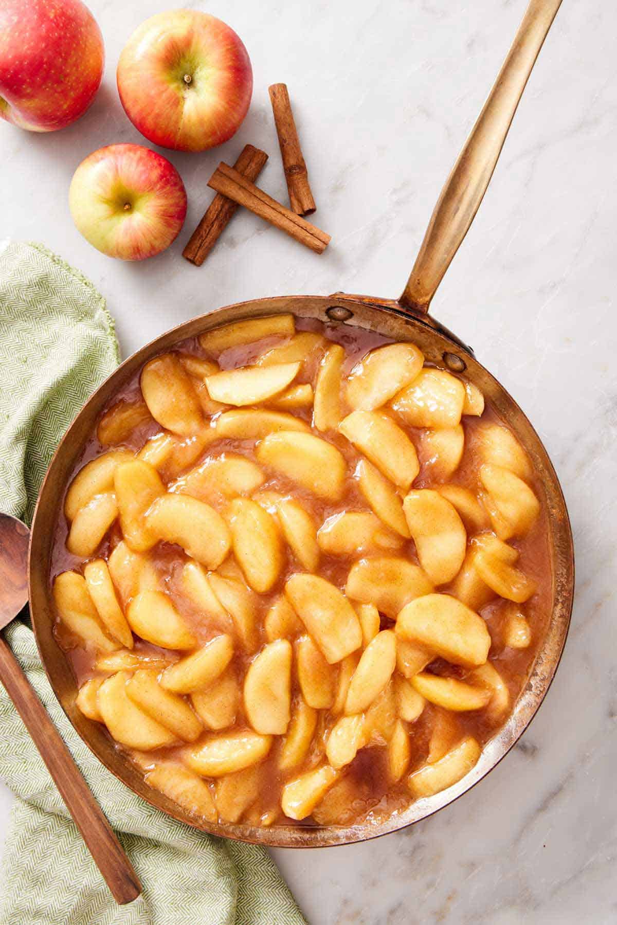Overhead view of a skillet of apple pie filling. Cinnamon sticks and apples on the side.