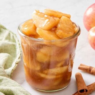 A jar of apple pie filling with cinnamon sticks and apples scattered around.