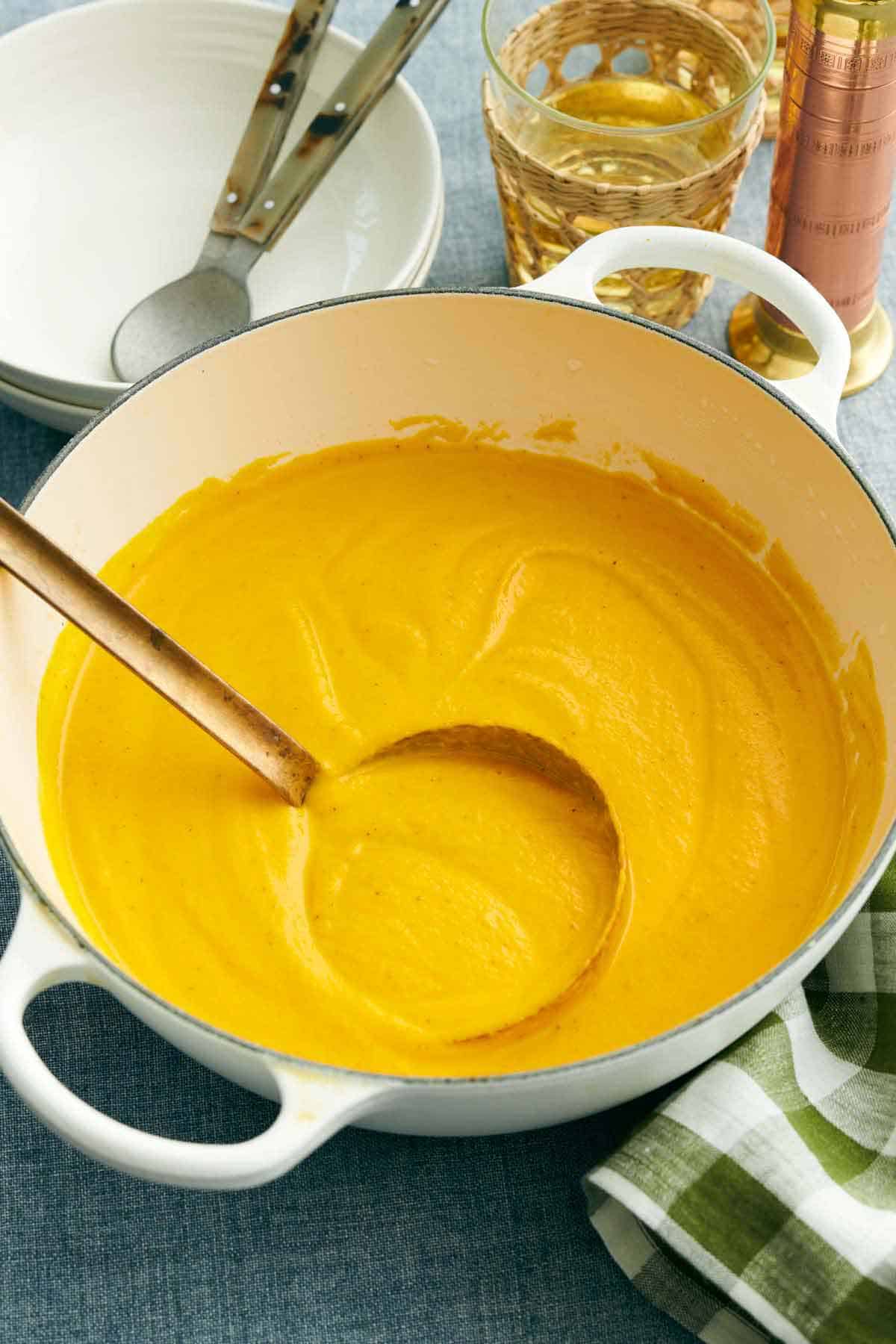A white Dutch oven of creamy carrot soup with a ladle. Bowls, spoons, and a drink in the background.