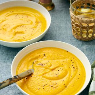 Pinterest graphic of a bowl of carrot soup with a spoon. A second bowl in the back along with drinks.