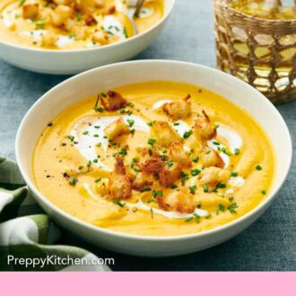 Pinterest graphic of a bowl of carrot soup with cream and croutons. A second bowl and a glass in the back.