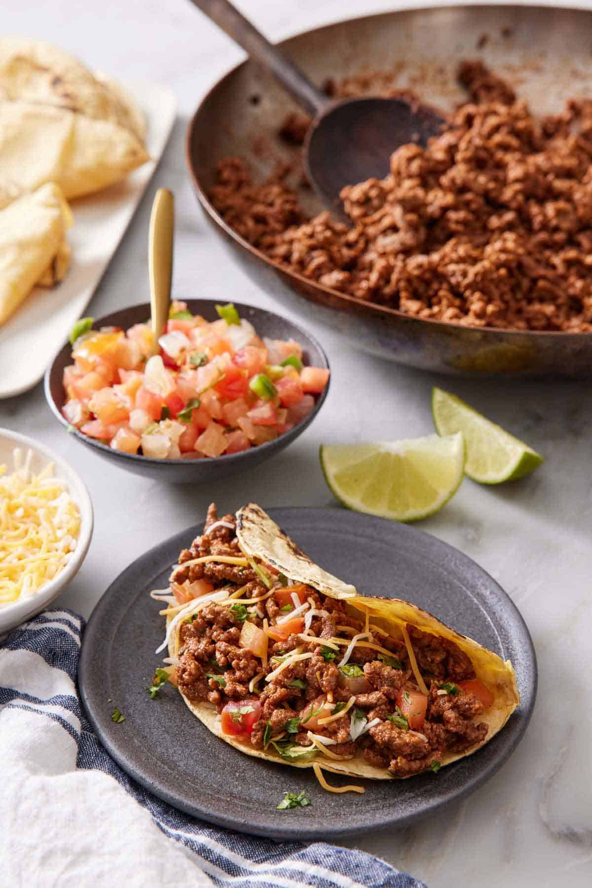 A plate with a ground beef taco topped with cheese, pico de gallo, and cilantro. A skillet with more ground beef, a bowl of pico de gallo, lime wedges, and tortillas in the background.