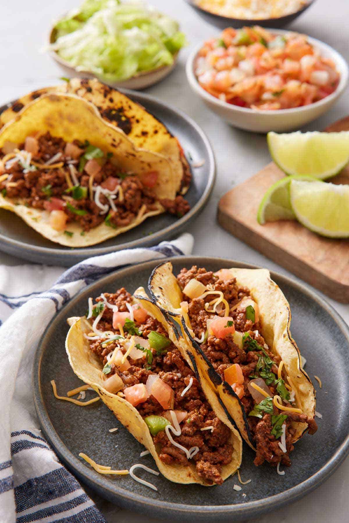 A plate with two ground beef tacos topped with cheese, pico de gallo, and cilantro. Another plate with two in the background along with bowls of toppings.