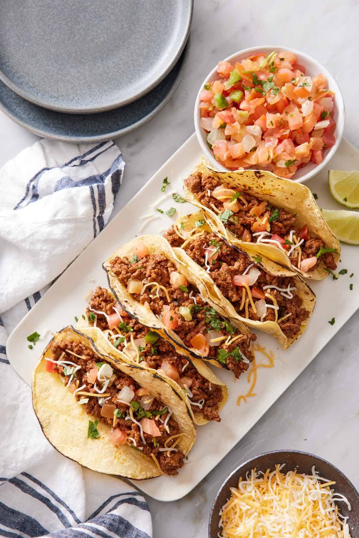 Overhead view of five ground beef tacos on a platter with a bowl of pico de gallo and lime wedges. A bowl of shredded cheese and some plates on the side.