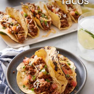 Pinterest graphic of two ground beef tacos topped with cheese, pico de gallo, and cilantro on a plate. More tacos in the background along with a drink.