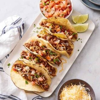 Overhead view of five ground beef tacos on a platter with a bowl of pico de gallo and lime wedges.