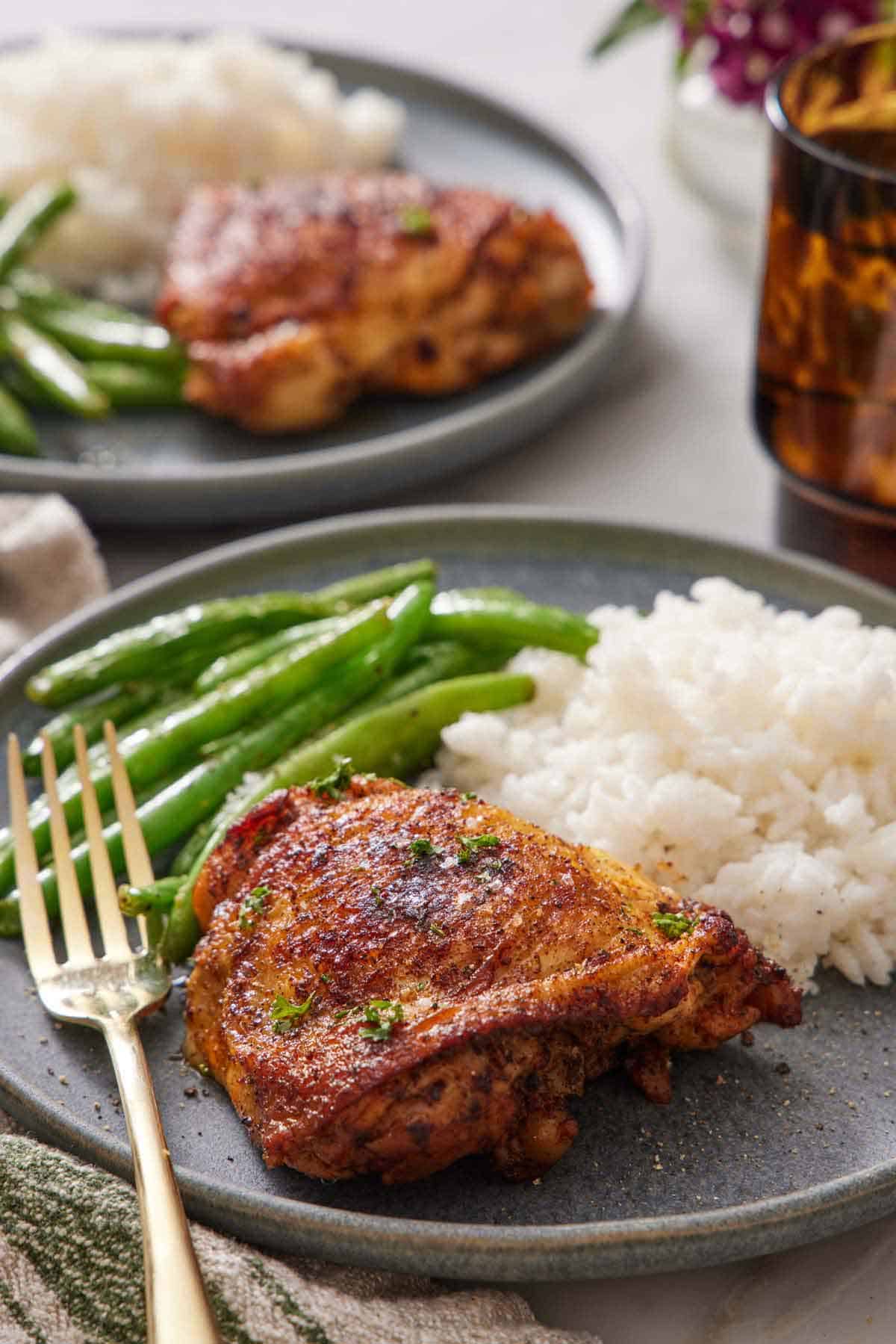 A close up view of an Instant Pot chicken thigh on a plate with rice and green beans. Another plated serving in the background.