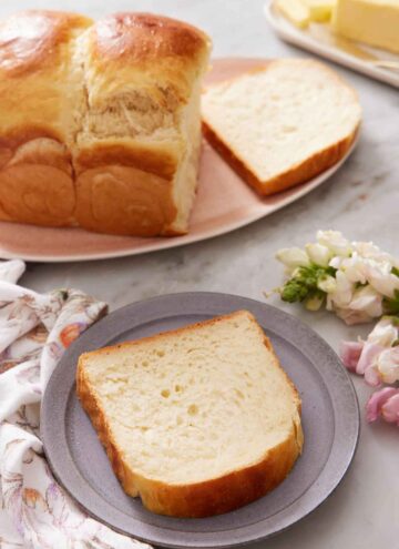 A plate with a slice of milk bread. The rest of the loaf in the back on a platter with one slice cut.