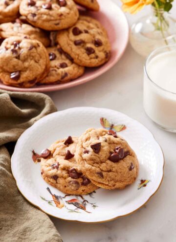 A plate with two pumpkin chocolate chip cookies with a glass of milk and platter of more cookies in the background.