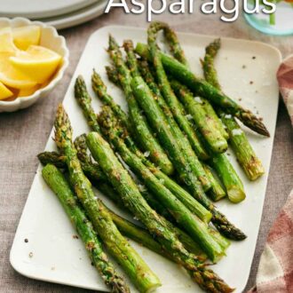 Pinterest graphic of a platter of roasted asparagus. Lemon wedges, stack of plates, and flowers in the background.