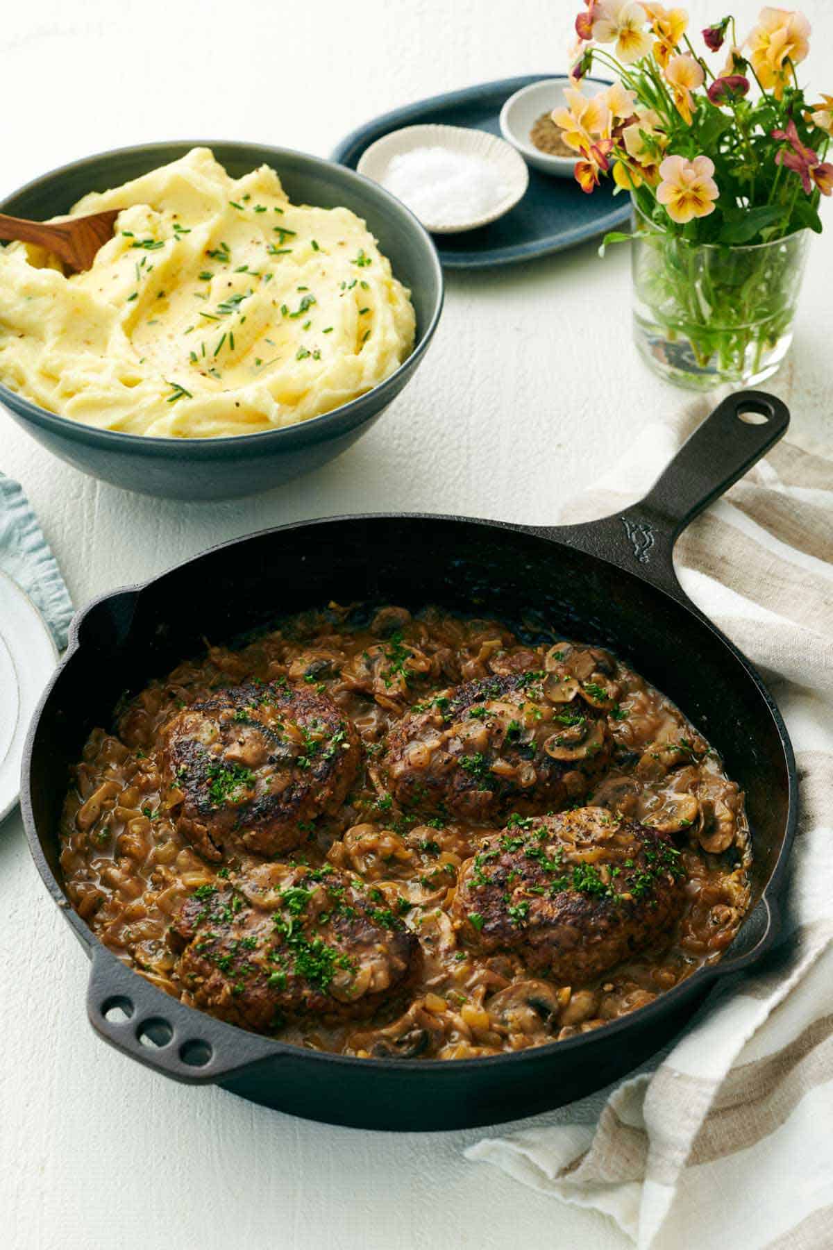A cast iron skillet of salisbury steaks with a bowl of mashed potatoes in the back along with a vase of flowers.