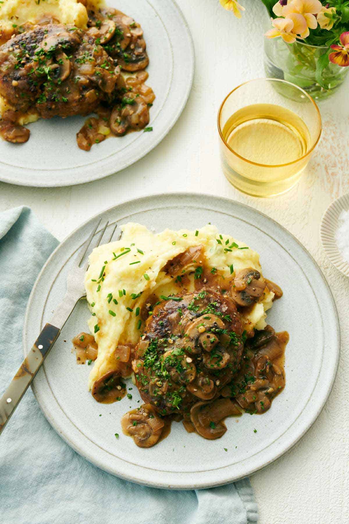 Overhead view of a plate with mashed potatoes and salisbury steak with a fork. A drink and another plated serving beside it.