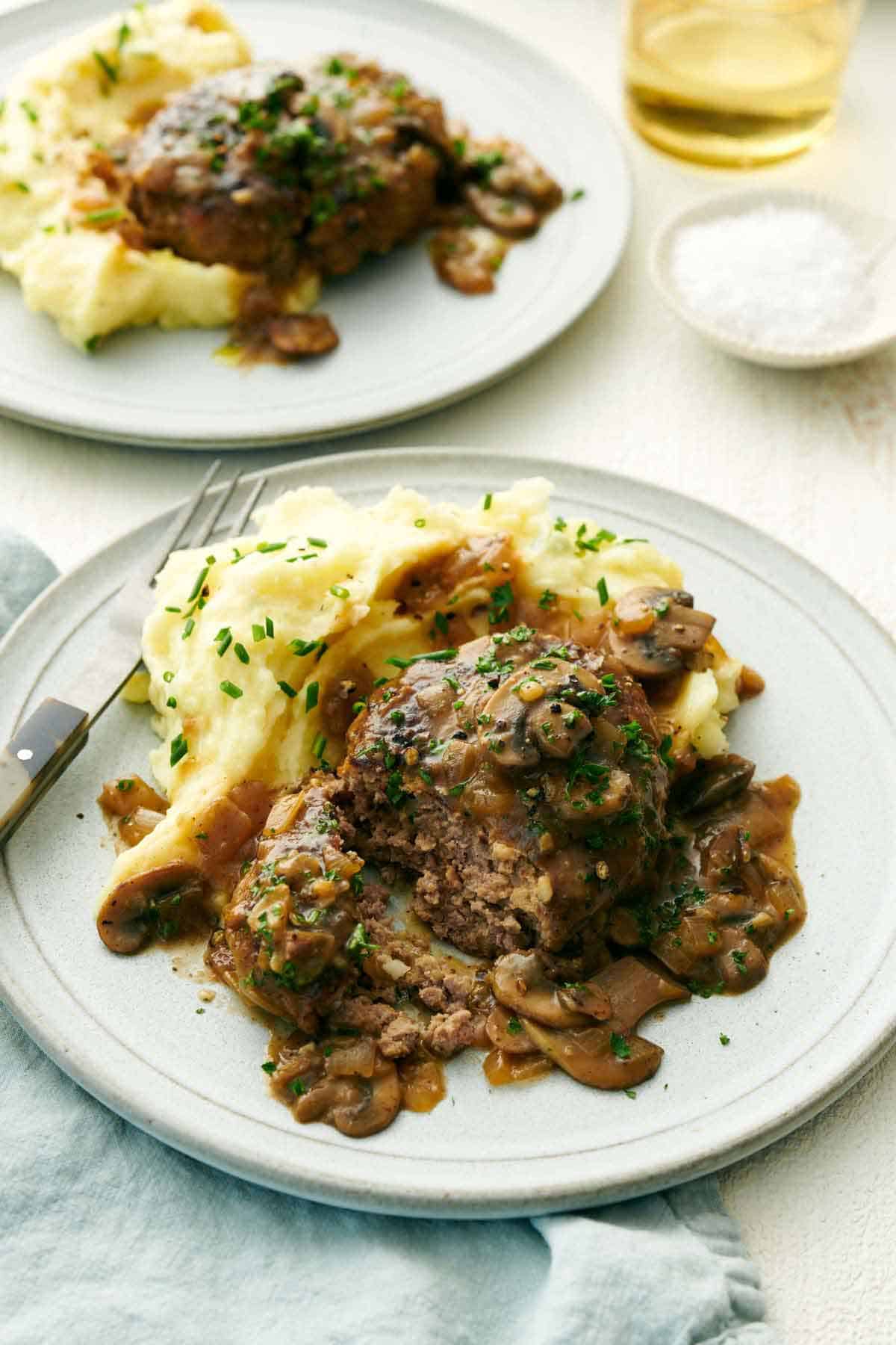 A plate of mashed potatoes and salisbury steak with the steak slightly cut opened. Another plated serving in the back.