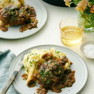 Pinterest graphic of a plate with salisbury steak and mashed potatoes. A second plated serving in the back with a drink, vase of flower, and skillet.