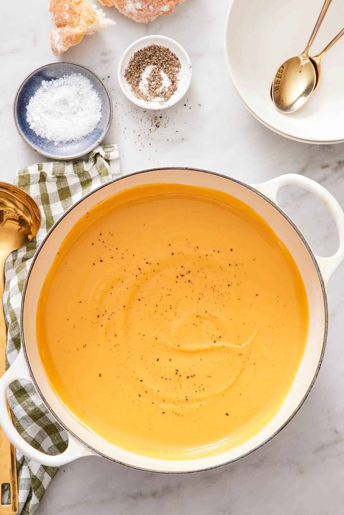 Overhead view of a pot of sweet potato soup topped with black pepper. A bowl of salt, pepper, stack of bowls, spoons, and bread on the side.