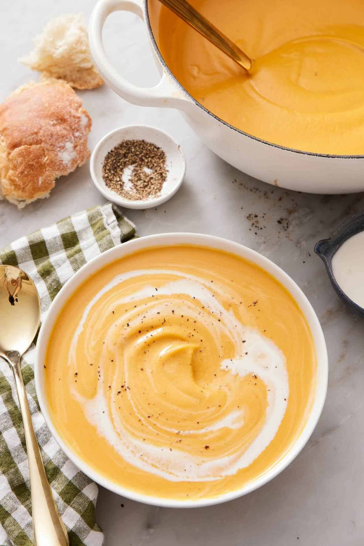 A bowl of sweet potato soup with cream swirled in topped with black pepper. A pot with more soup in the back along with pepper and torn bread.