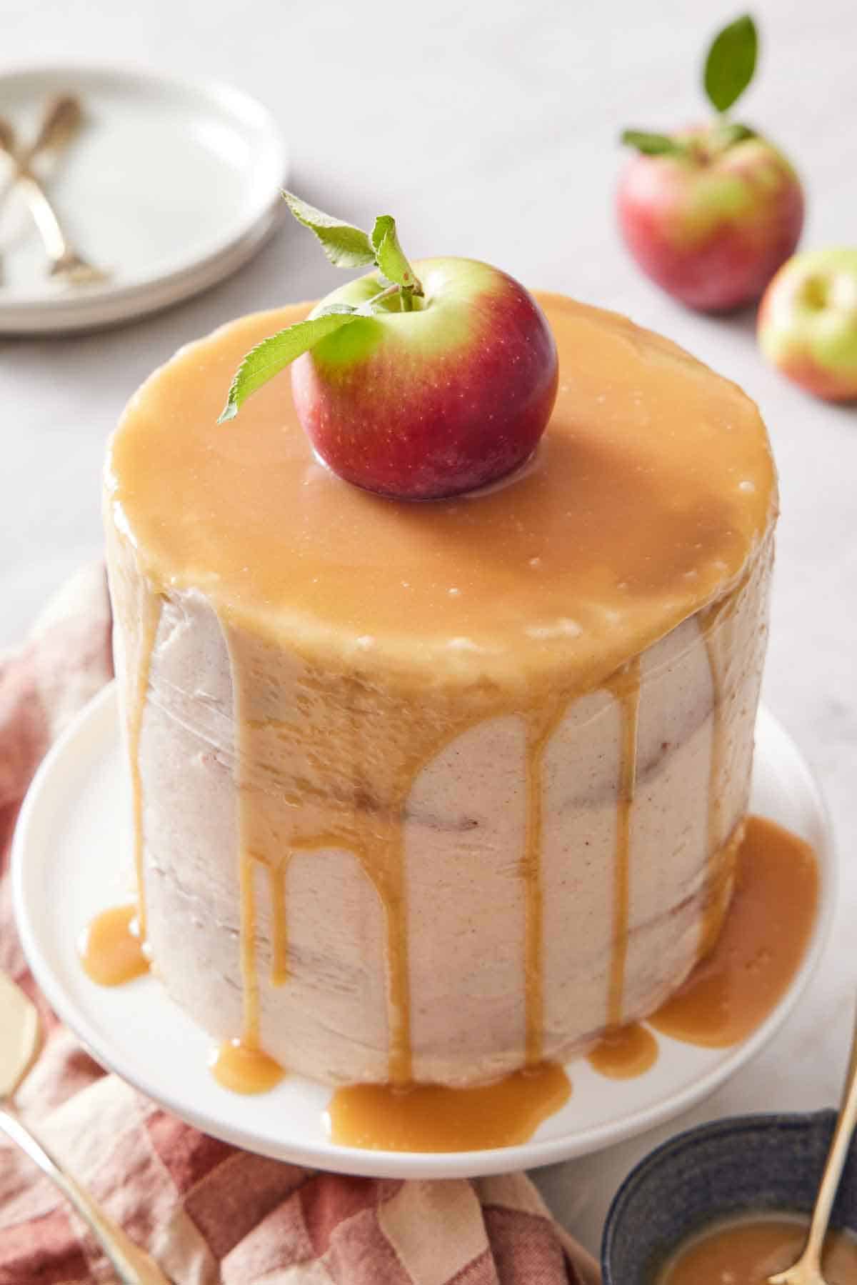 A caramel apple cake topped with an apple with caramel dripping down the sides on a white cake stand.