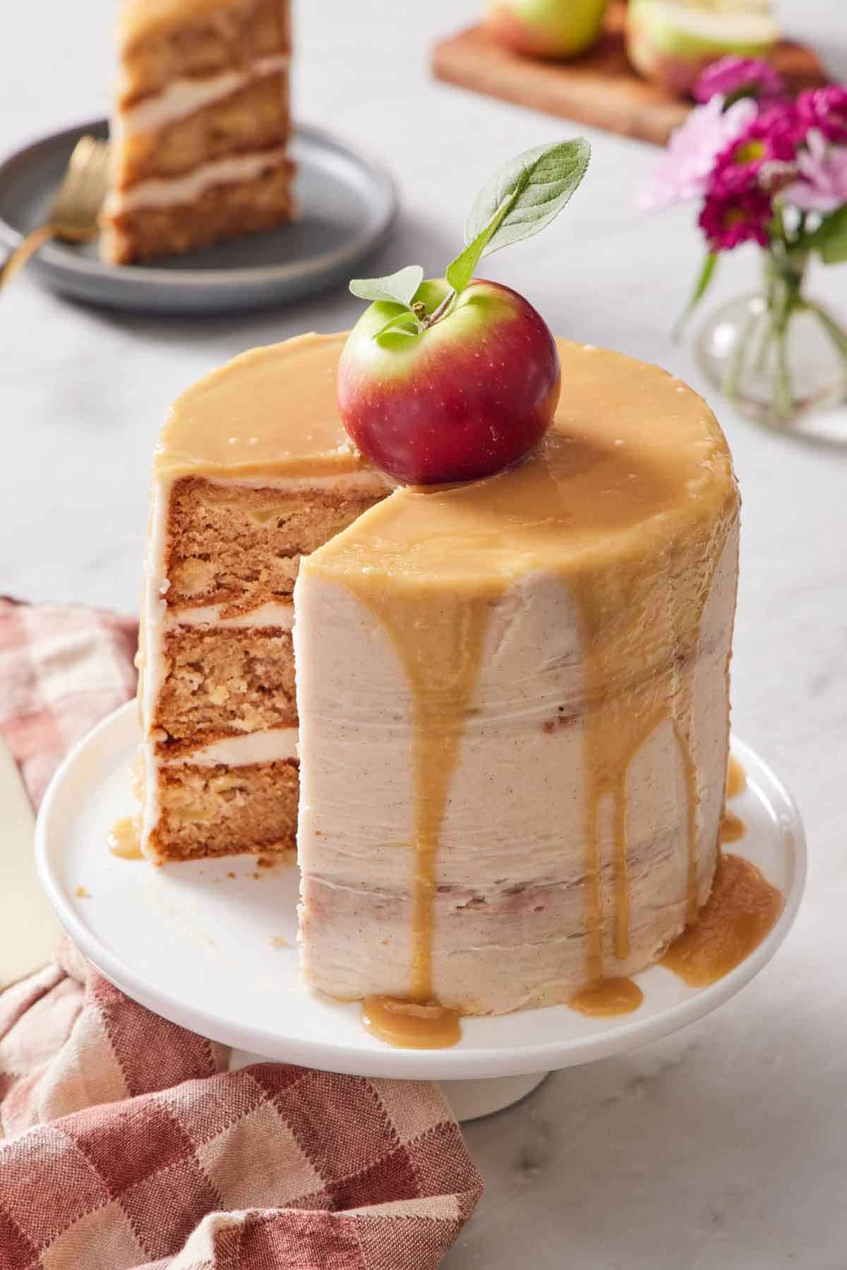 A caramel apple cake with a slice cut out on a white cake stand. An apple on top and caramel dripping down the sides of the cake. A slice on a plate in the background.