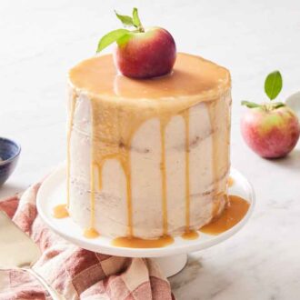 A caramel apple cake on a white cake stand. An apple on top and caramel dripping down the sides of the cake. Apples, a bowl of caramel, and stack of plates with forks in the background.