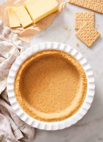 Overhead view of a pie dish containing a graham cracker crust. Butter and graham crackers in the background.